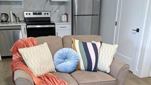 A living room with a sofa, pillows, a blanket, a wicker basket, and a kitchen in the background. - Steps to Beach • 2BR White Rock Suite • Sleeps 4 (Surrey)