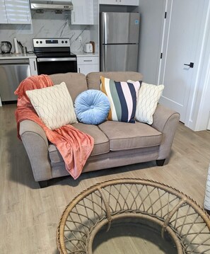 A living room with a sofa, pillows, a blanket, a wicker basket, and a kitchen in the background. - Steps to Beach • 2BR White Rock Suite • Sleeps 4 (Surrey)