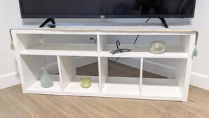 A white TV stand with a flat-screen TV on top, three shelves, and a small decorative item on the middle shelf. - Steps to Beach • 2BR White Rock Suite • Sleeps 4 (Surrey)