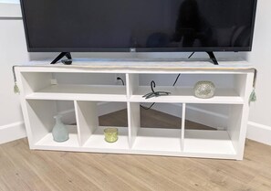 A white TV stand with a flat-screen TV on top, three shelves, and a small decorative item on the middle shelf. - Steps to Beach • 2BR White Rock Suite • Sleeps 4 (Surrey)