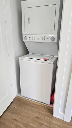 Une petite buanderie blanche, équipée d’un lave-linge et d’un sèche-linge, avec un plancher en bois et une porte donnant sur une autre pièce.