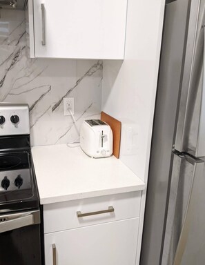 A kitchen with white cabinets, a toaster, a stove, and a refrigerator. - Steps to Beach • 2BR White Rock Suite • Sleeps 4 (Surrey)