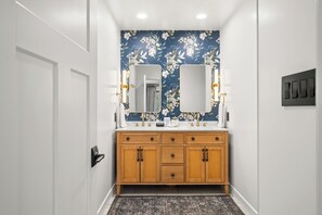 A bathroom with a floral wallpaper, a white door, a wooden vanity with drawers, a mirror, and a towel rack. - Cozy 2-bedroom cottage in serene Brown County Indiana (Nashville)