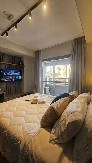 A hotel room with a large bed, a television mounted on the wall, and a view of the city through the window. - Condominio Hub Brooklin (São Paulo)