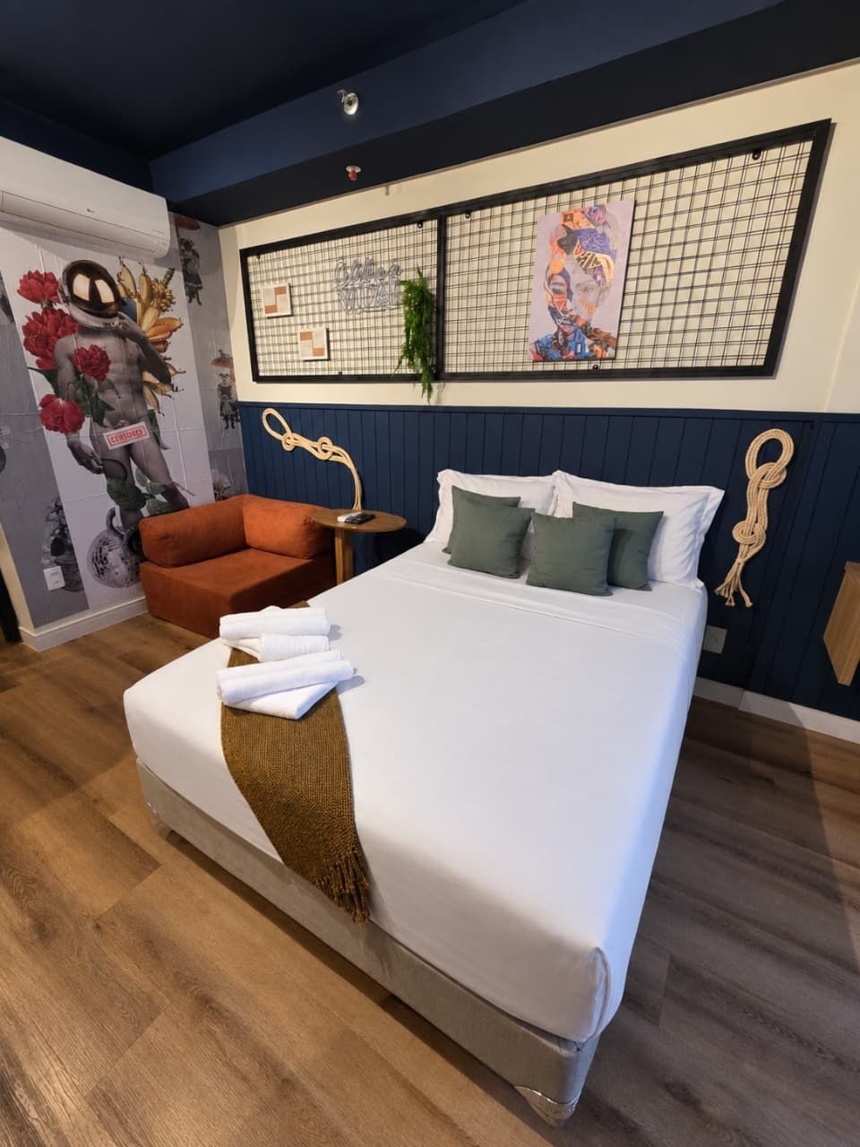 Ein ordentlich bezogenes Bett mit weißen Leinen und grünen Kissen, ein kleiner orangefarbener Sessel und eine Wand mit Rastern und Kunstwerken.
