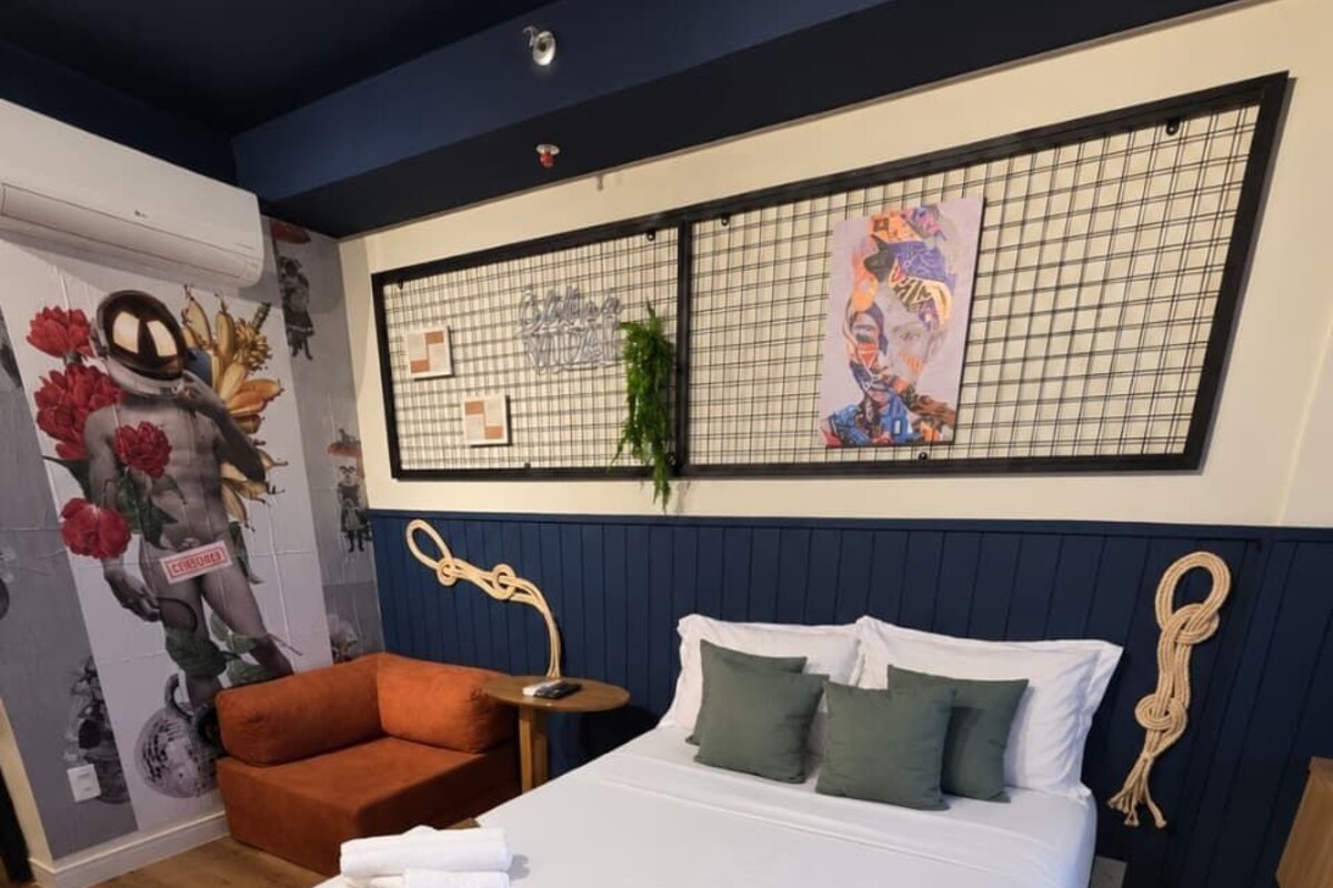 A neatly made bed with white linens and green pillows, a small orange chair, and a wall with a grid pattern and artwork.