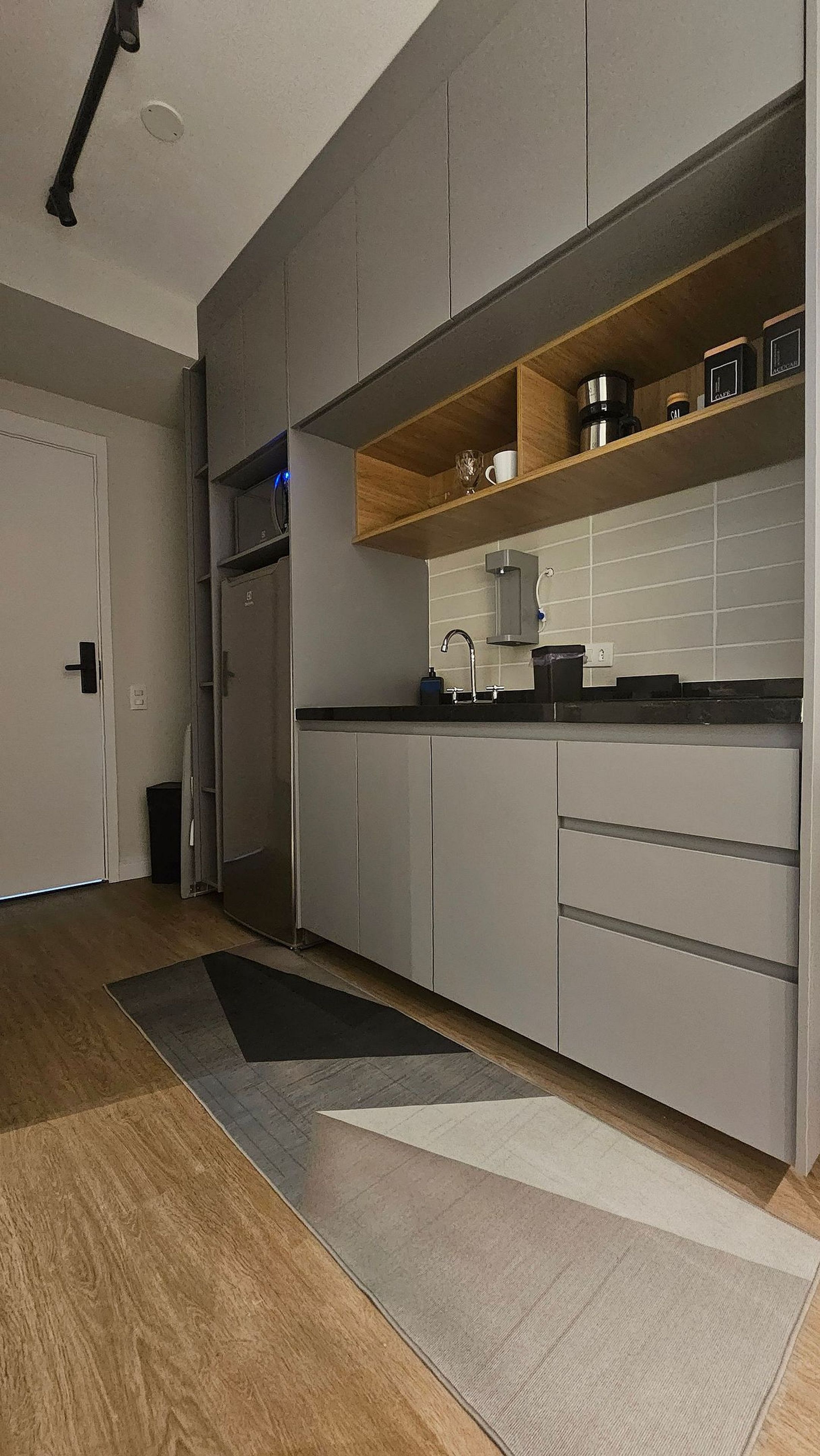 Une cuisine moderne avec des meubles gris, un plan de travail noir et une étagère en bois au-dessus de l’évier.