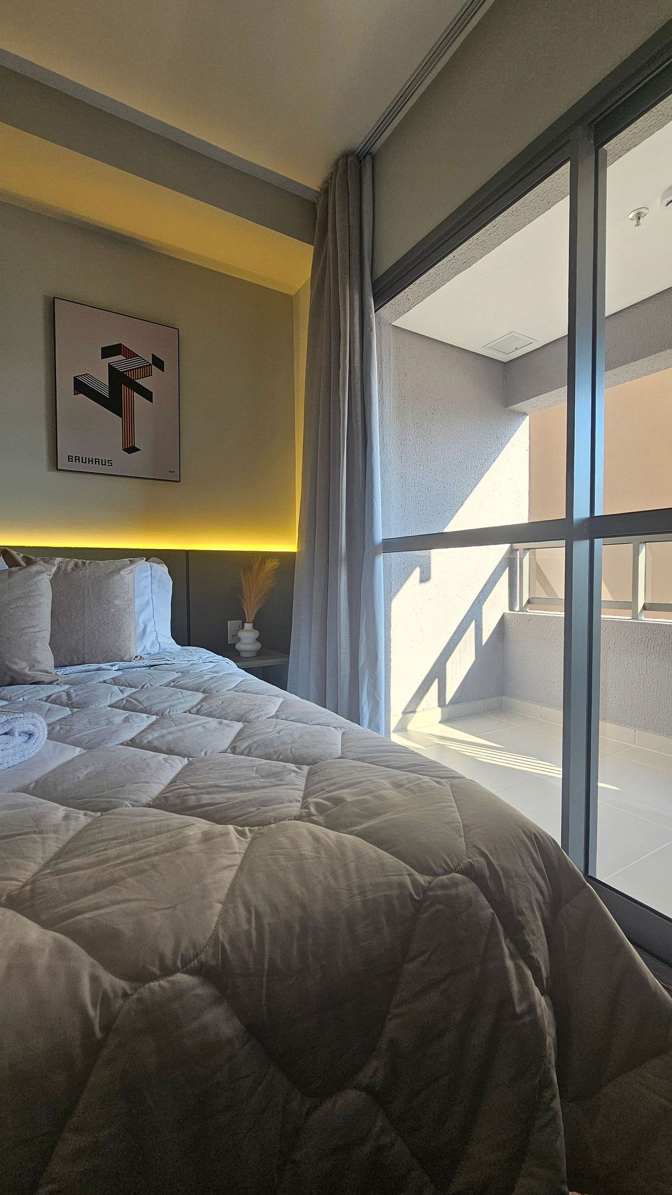 A hotel room with a large bed, a nightstand, a framed picture on the wall, and a view of the outside balcony.