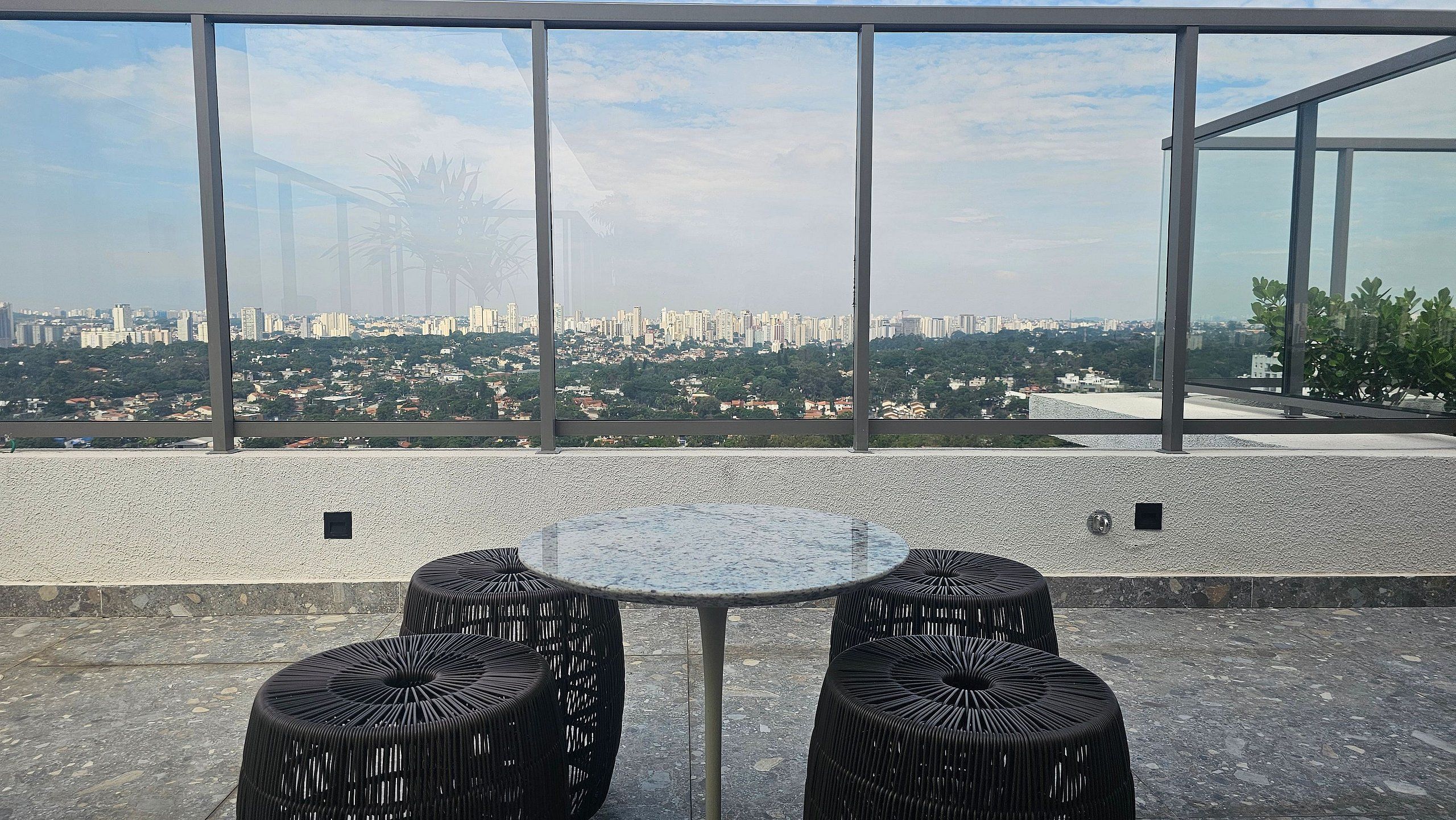 A terrace with a glass table and two wicker chairs, overlooking a cityscape.