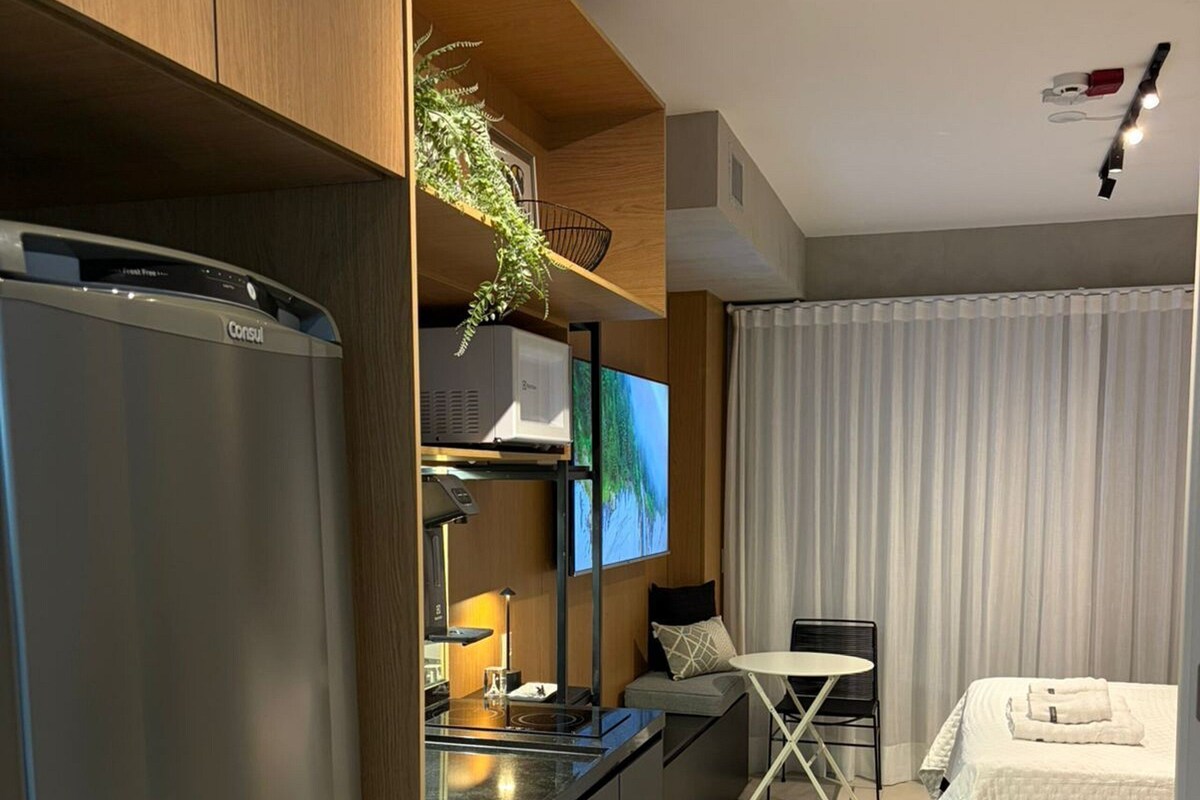 A modern hotel room with a kitchenette, a flat-screen TV, a bed with a white duvet, and a dining area with a table and chairs.