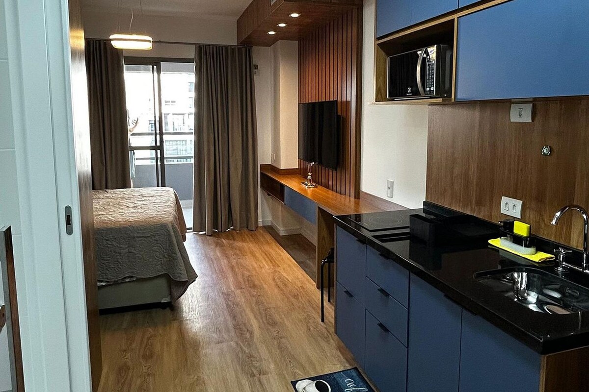 A compact hotel room with a kitchenette, a bed with a view, and a wooden floor.