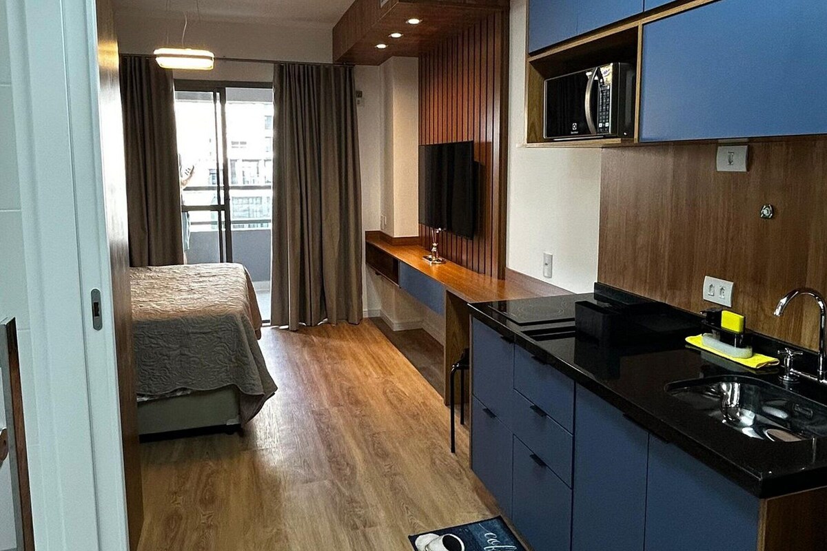 A compact hotel room with a kitchenette, a bed with a view, and a wooden floor.