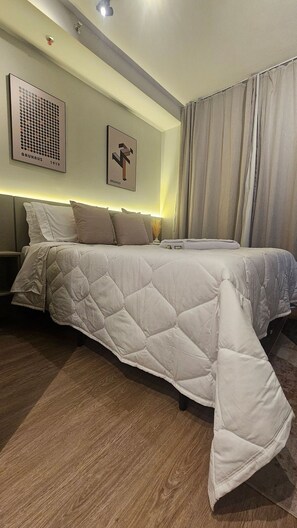 A modern bedroom with a large bed, a headboard, a wall-mounted artwork, and a window with curtains. - Condominio Hub Brooklin (São Paulo)