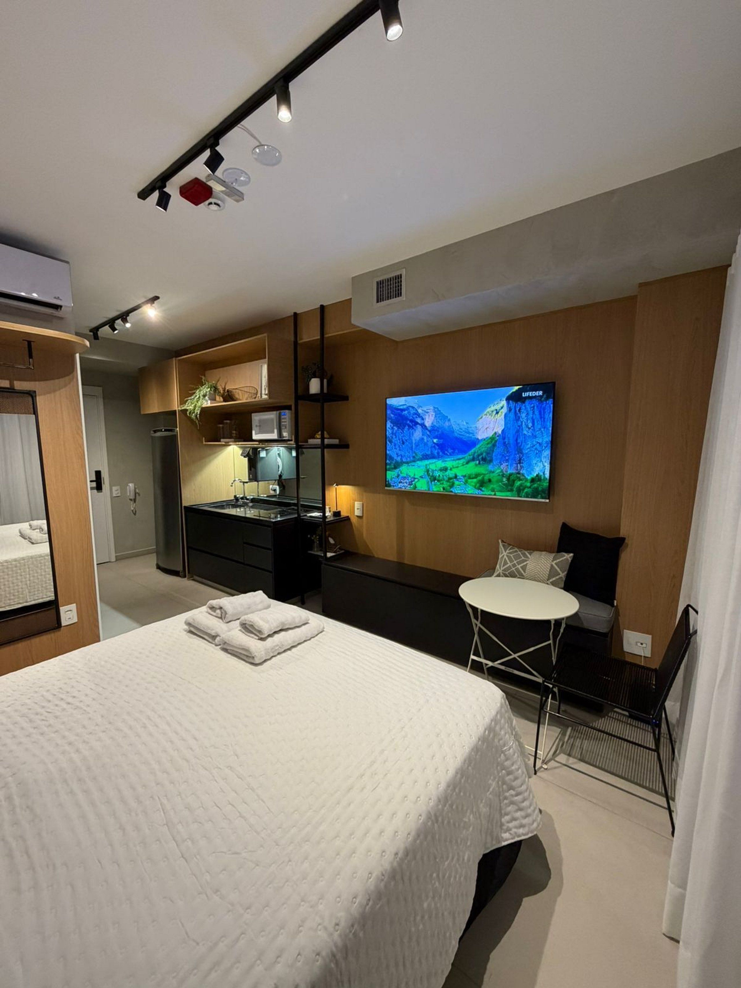 Ett modernt hotellrum med en stor säng, en tv som är monterad på väggen, ett litet bord och en liten köksdel.