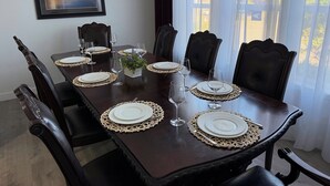 Mesa de comedor con platos blancos, cubiertos y vasos, rodeada de sillas de madera oscura.