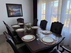 A dining table set with white plates, silverware, and glassware, surrounded by dark wooden chairs.
