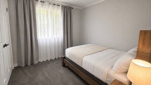 Un dormitorio con cama, mesita de noche con lámpara, una puerta y una ventana con cortinas.