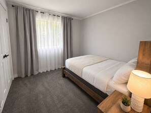 A bedroom with a bed, a bedside table with a lamp, a door, and a window with curtains.