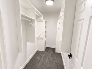 En smal genomgångsklädkammare med vita hyllor och plats för att hänga kläder, ett grått mattgolv och en vit dörr längst in.