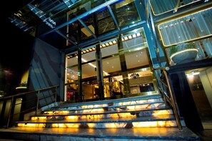 Fachada de hotel de vidrio, una escalera que conduce a la entrada y una planta en maceta a la derecha.