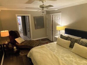 A hotel room with a bed, a ceiling fan, a nightstand, a lamp, and a door leading to another room. - Warm & Cozy Condo Feels Like Home (Dallas)