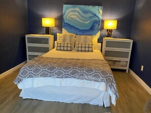 A bedroom with a bed, two bedside tables, a nightstand, a wall-mounted lamp, and a large abstract painting. - Spacious 2BR Condo 208 (Dallas)