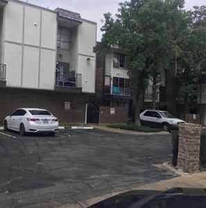 A parking lot with a white car and a white SUV. There are buildings with balconies and a tree in the background. - Spacious 2BR Condo 208 (Dallas)