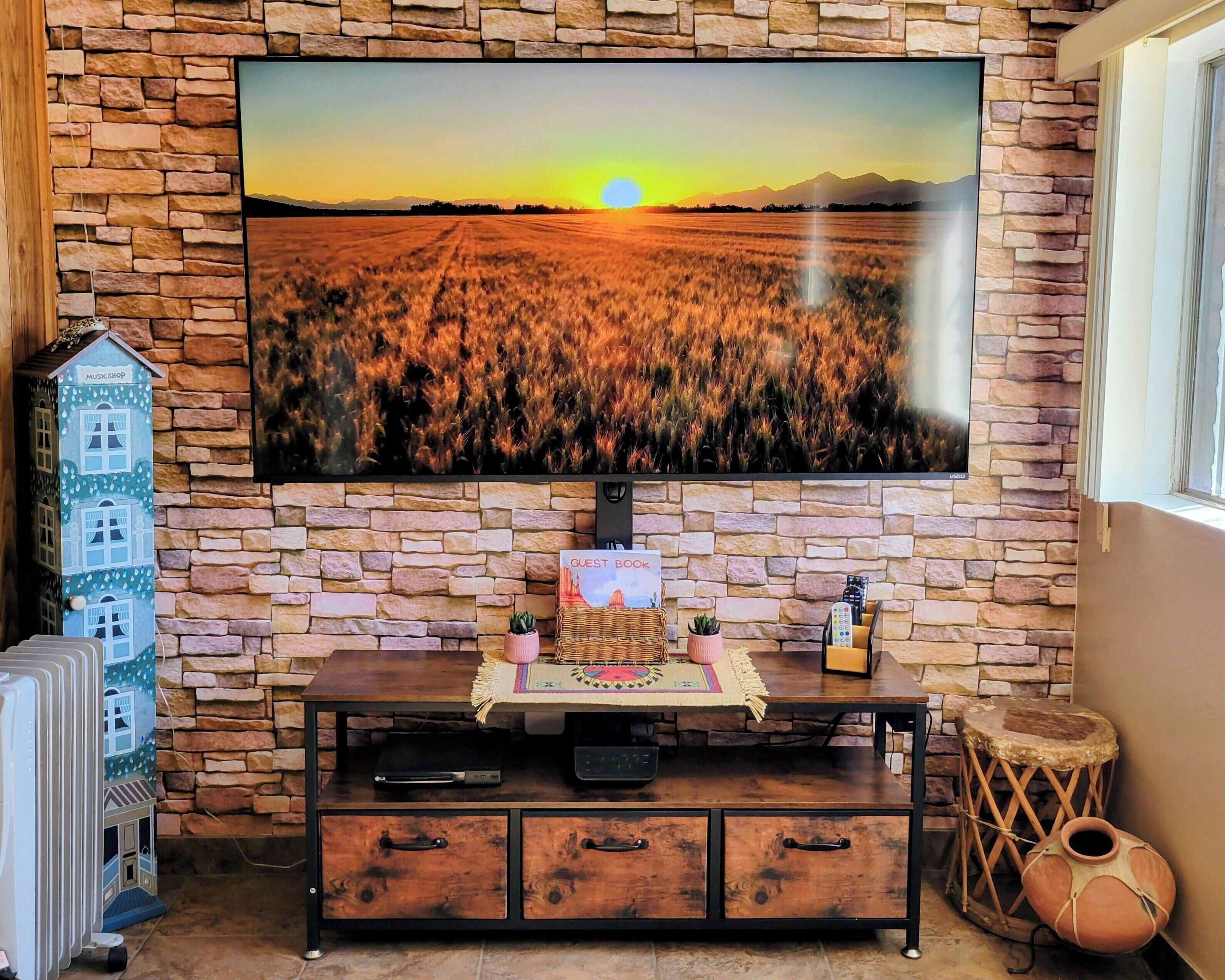Una habitación con pared de piedra, un televisor de pantalla plana mostrando un atardecer, un mueble de madera con un libro de visitas, y una mesita con un control remoto.