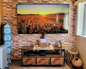 A room with a stone wall, a flat-screen TV displaying a sunset, a wooden cabinet with a guest book, and a small table with a remote control. - Lovely 2-bedroom house in the heart of Yuma - Pet Friendly! (Yuma)