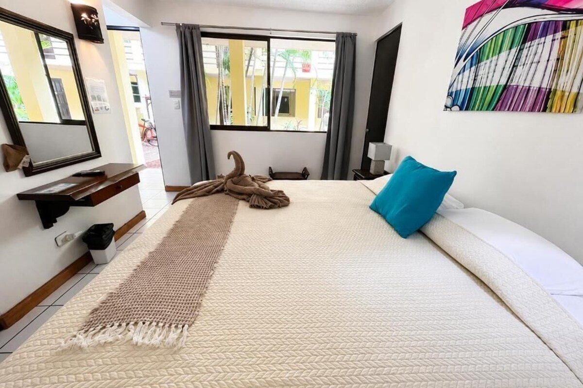 Habitación de hotel con cama, un ventanal grande, un espejo, una mesita de noche y un cuadro abstracto de colores en la pared.