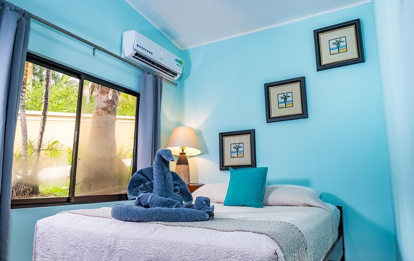 A hotel room with a bed, a window with curtains, an air conditioning unit, a lamp, and framed pictures on the wall.