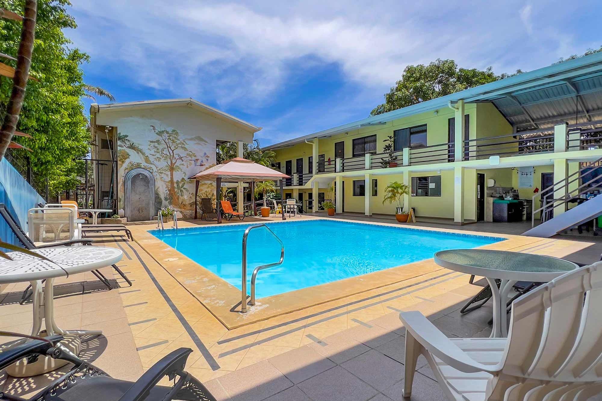 Una piscina con sillones y mesas, rodeada de edificios con balcones y un mural.