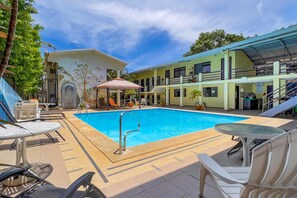 Una piscina con sillones y mesas, rodeada de edificios con balcones y un mural.