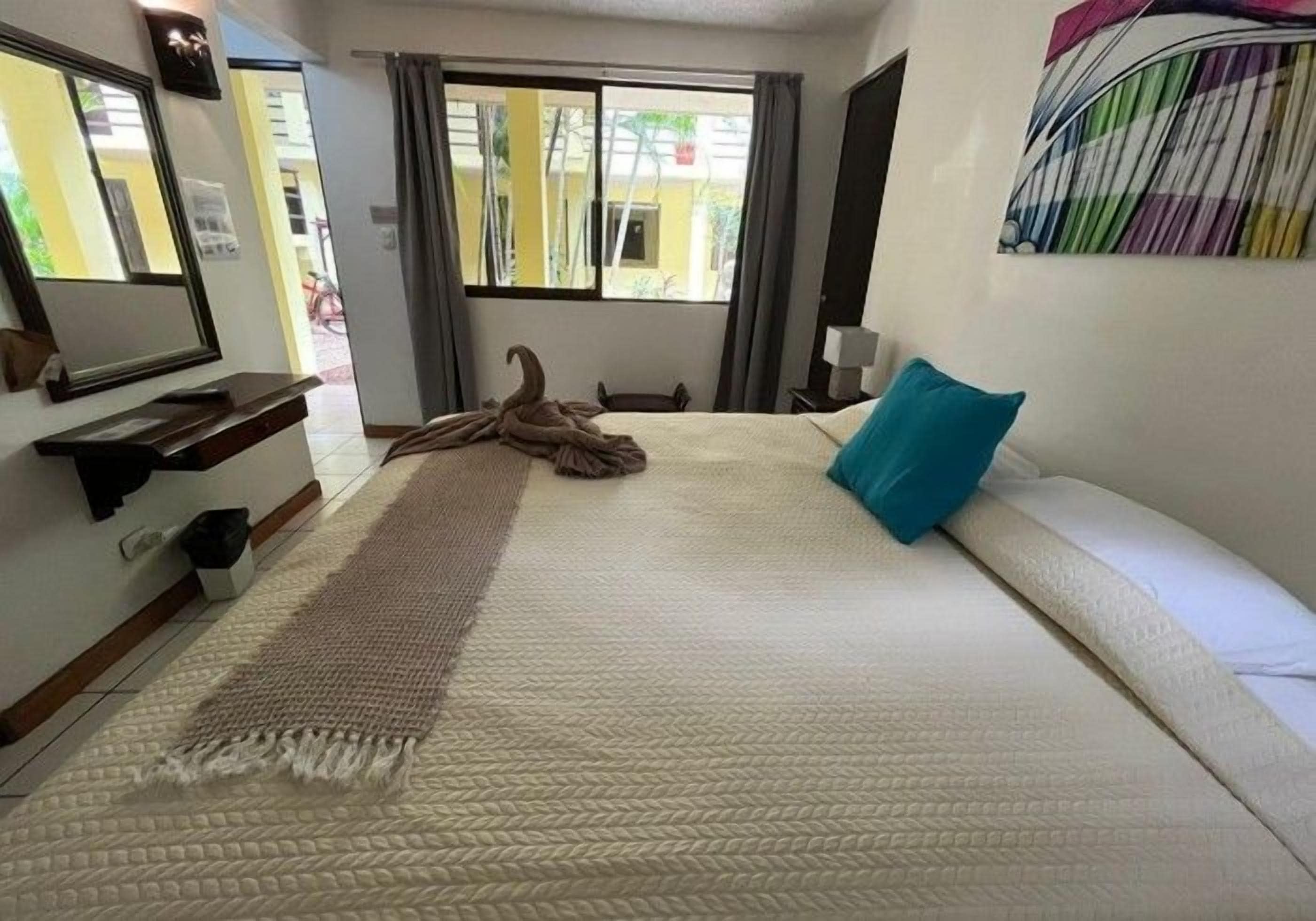 Habitación de hotel con cama, ventana grande, espejo, mesita de noche y una obra de arte colgada en la pared.