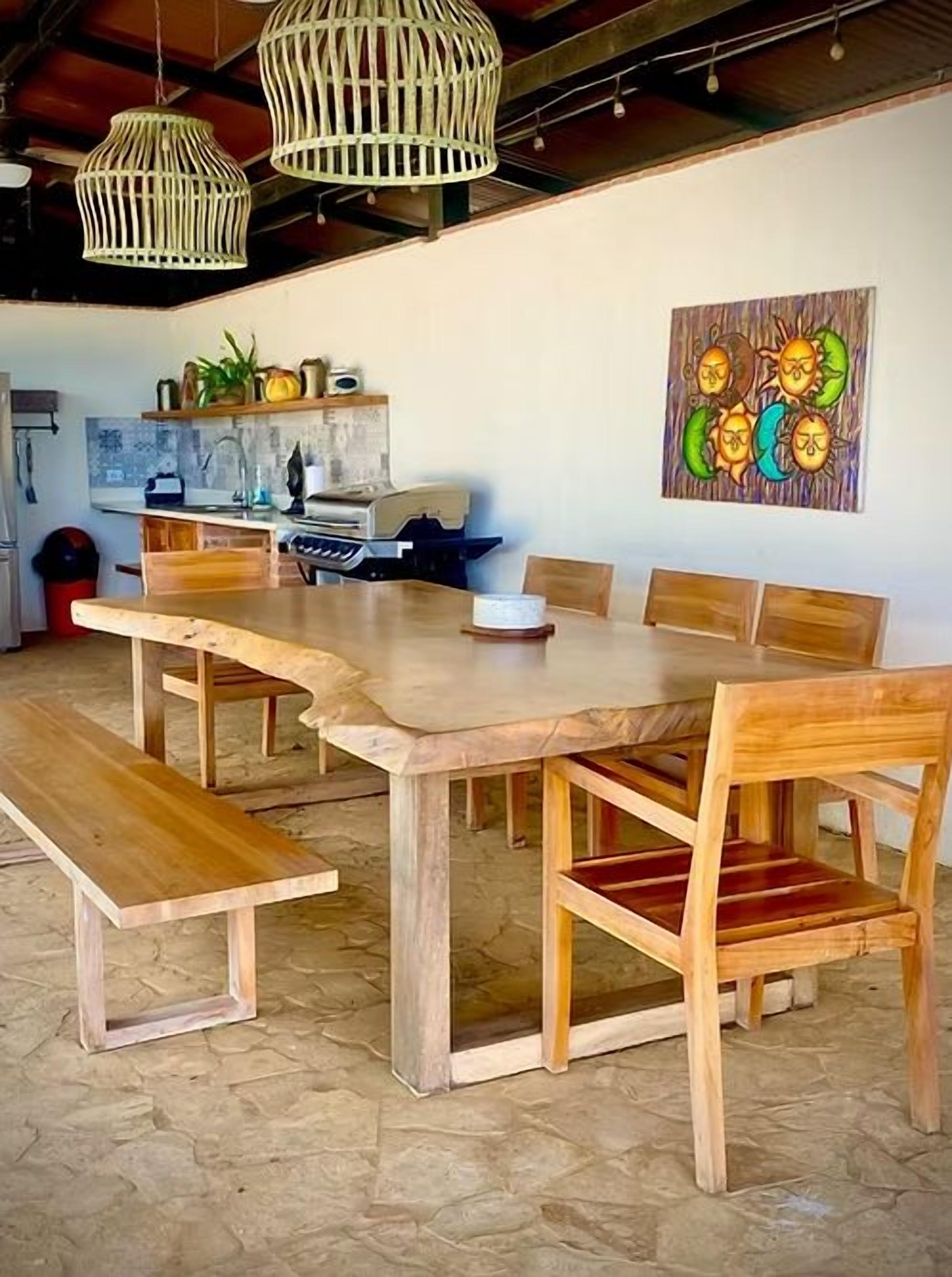 Un comedor con una larga mesa de madera y bancos, una cocina al fondo, y un muro decorado con una obra de arte colorida.