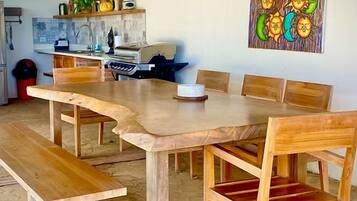 Un comedor con una larga mesa de madera y bancos, una cocina al fondo, y un muro decorado con una obra de arte colorida.