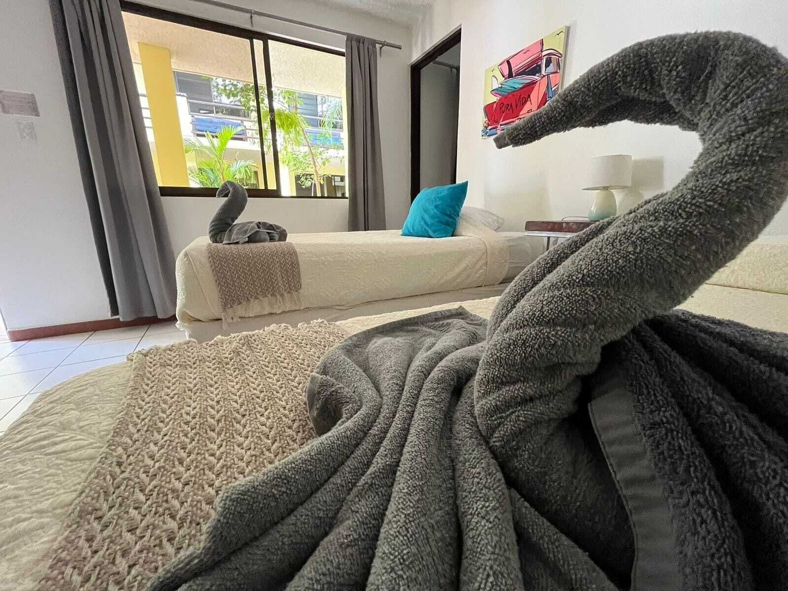 Ein Zimmer mit einer großen, kuscheligen, grauen, schwanenförmigen Dekoration auf dem Bett, einem Sofa mit einem blauen Kissen und einem Fenster mit Blick auf Gebäude und Grünflächen.