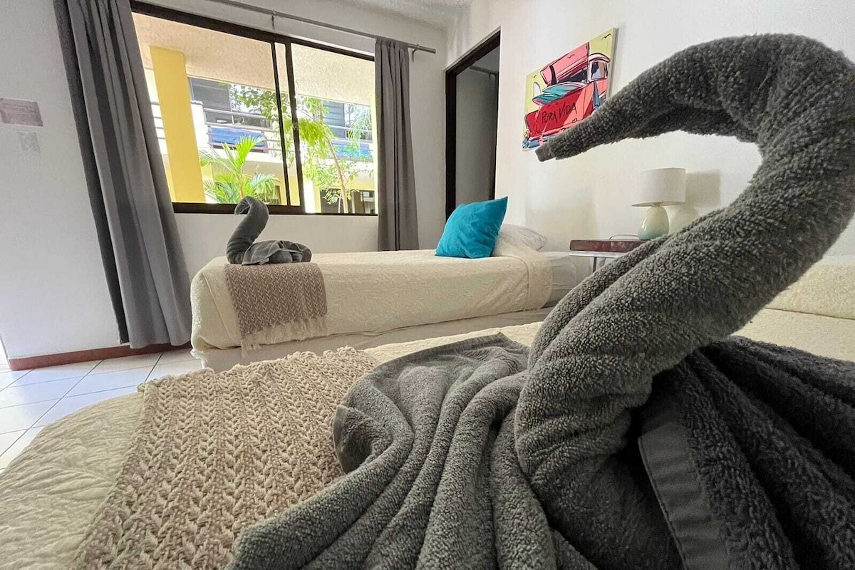 Una habitación con una decoración grande y mullida con forma de cisne en un cama, un sofá con un cojín azul y una ventana con vistas a edificios y follaje.