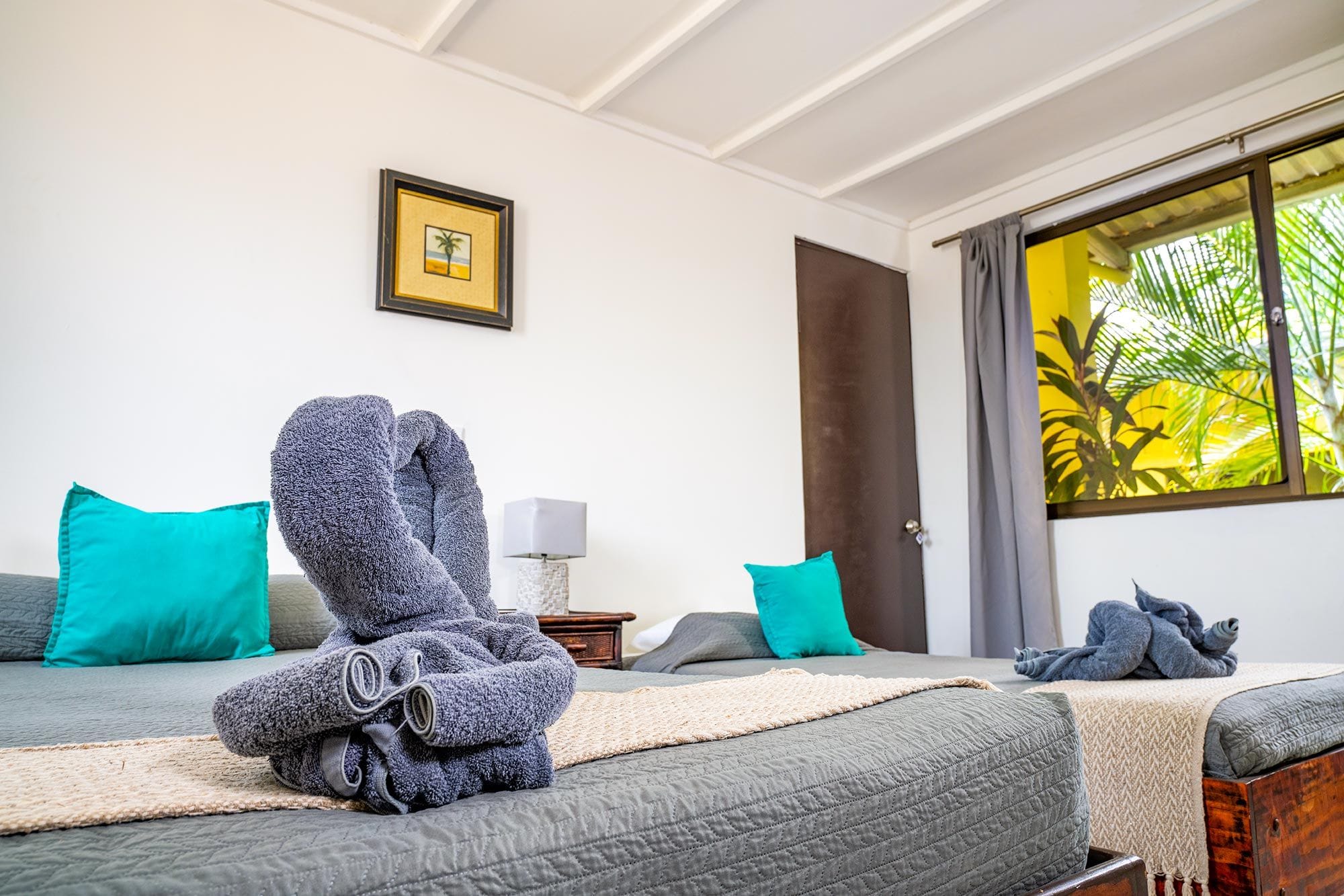 Ein Hotelzimmer mit Bett, Kissen, einem zusammengerollten Handtuch, einem gerahmten Bild und einem Fenster mit Blick auf Grünflächen.