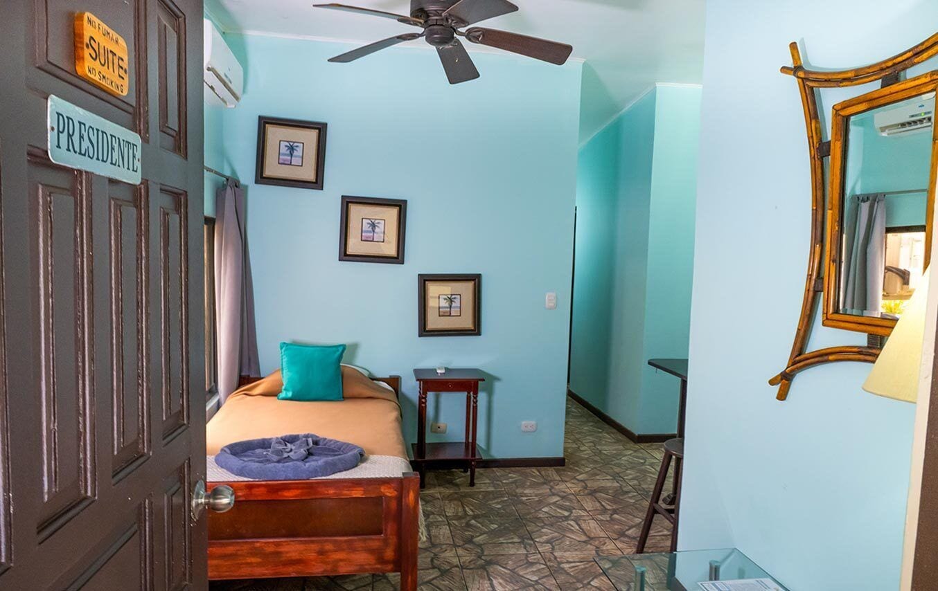 A room with a bed, a ceiling fan, framed pictures on the wall, and a door labeled 'Presidente.'