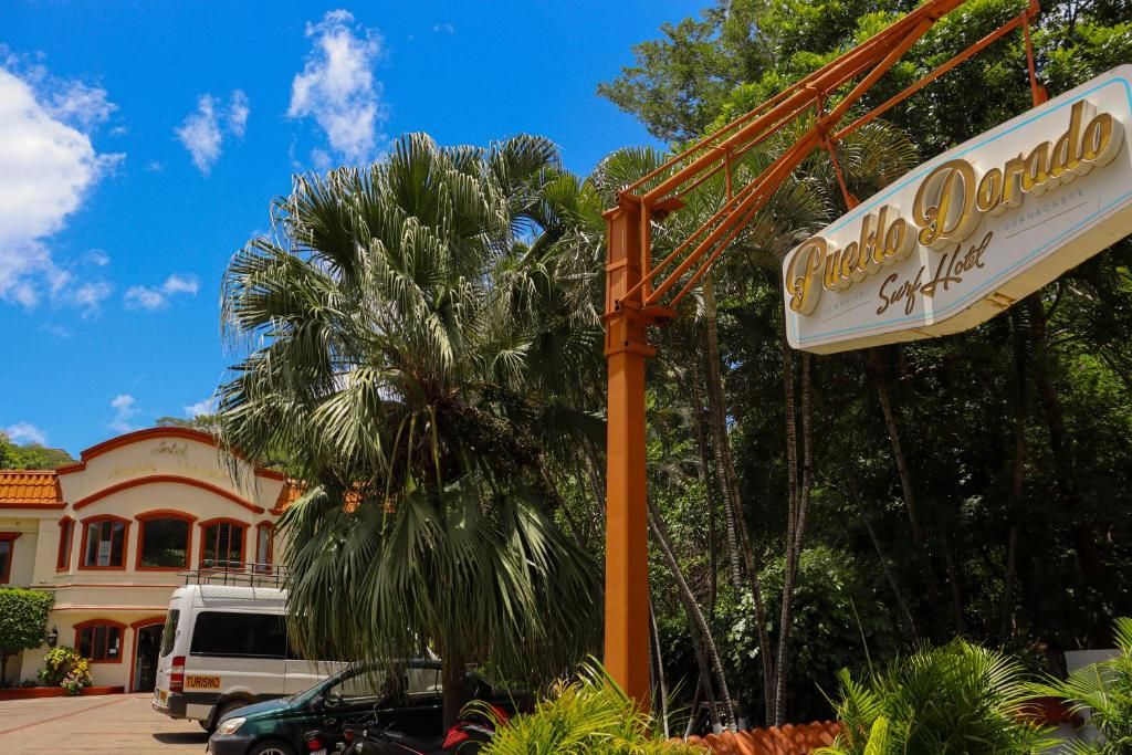 A hotel entrance with a sign reading 'Pueblo Dorado Surf Hotel,' palm trees, and a building with a red and white facade.