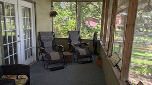 A covered porch with two reclining chairs, a small table, and a window with a view of a red building and greenery.