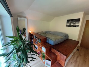 Une chambre à coucher de taille modeste, avec un lit en mezzanine, une bibliothèque en bois, une grande plante en pot et un tableau encadré accroché au mur.