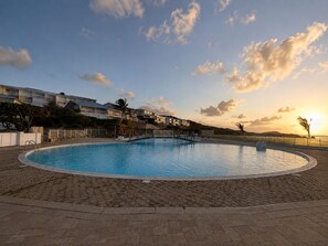 A swimming pool with a paved deck, surrounded by a fence, and buildings in the background. - The Ocean Pearl Studio - Ocean View & Beach Access (Orient Bay)