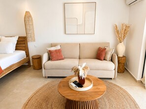 A modern living room with a sofa, a round wooden coffee table, a hanging light fixture, and a wall-mounted abstract art piece. - The Ocean Pearl Studio - Ocean View & Beach Access (Orient Bay)