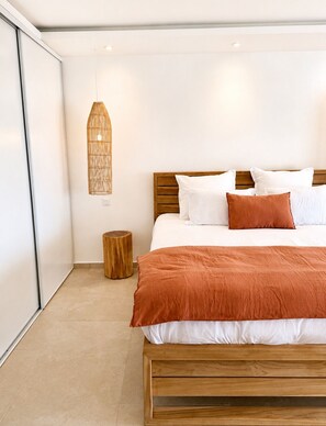 A bedroom with a bed, pillows, a bedside table, a hanging light fixture, and a wooden stool. - The Ocean Pearl Studio - Ocean View & Beach Access (Orient Bay)
