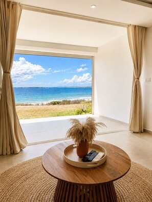 A room with a large window overlooking a beach, a round wooden table with a potted plant, and beige curtains. - The Ocean Pearl Studio - Ocean View & Beach Access (Orient Bay)