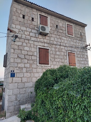 A stone building with three windows, each with wooden shutters, and an air conditioning unit mounted on the wall.
