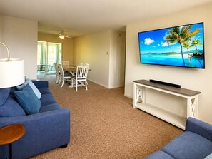 A living room with a blue sofa, a TV mounted on the wall, and a dining area in the background. - Fort Myers Monthly Rental — Sunroom, Utilities Included (Fort Myers)