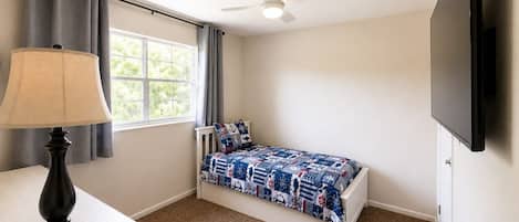 A bedroom with a single bed, a desk, a lamp, a ceiling fan, and a TV.